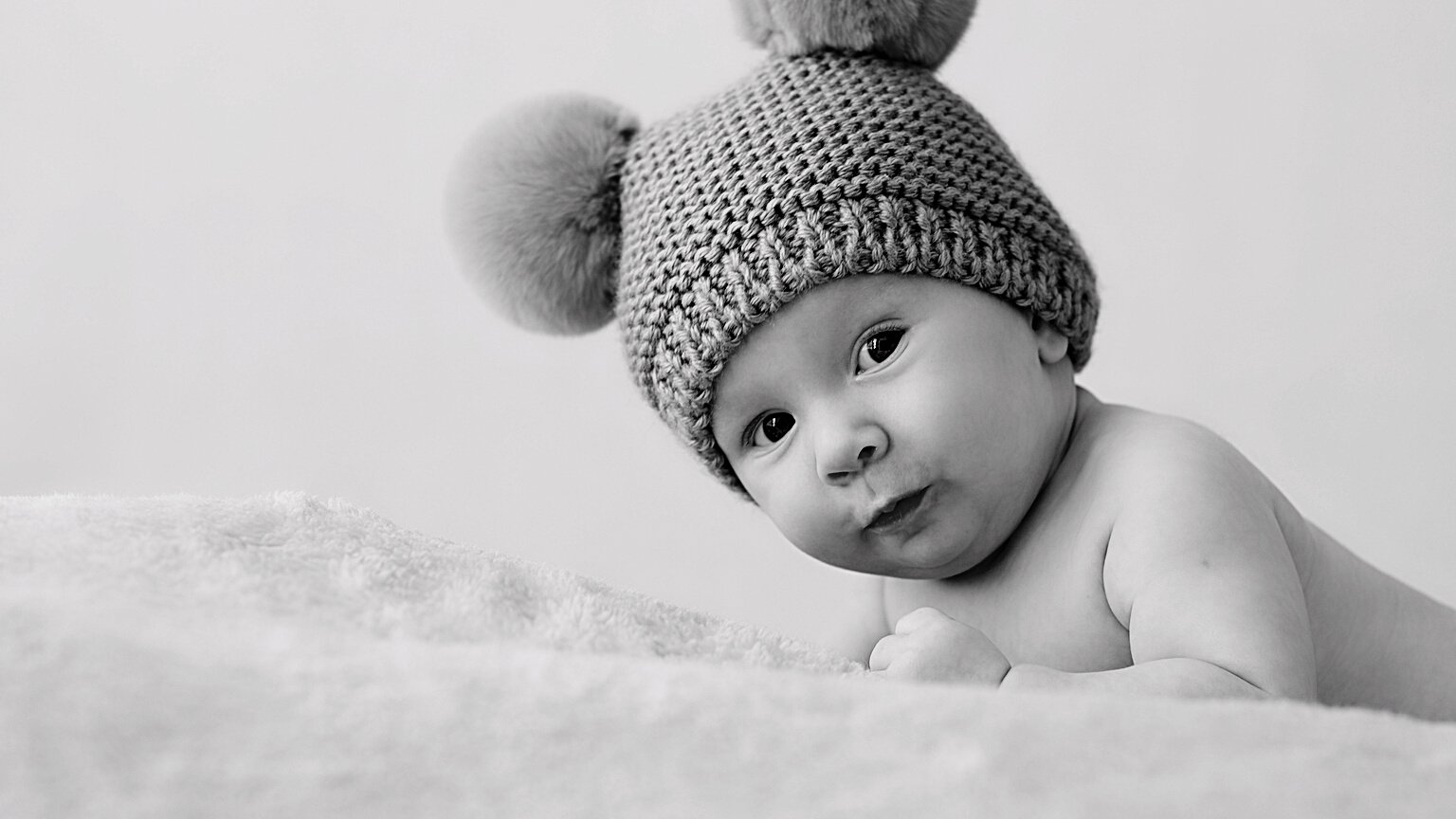 A Happy Newborn Baby in a Blue Hat with Pompoms Looks into the C