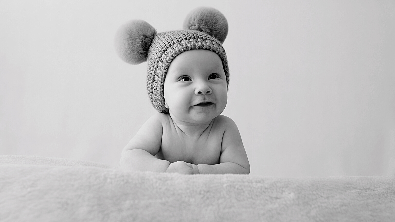 A Smiling Infant Baby in a Blue Hat with Pompoms Looks into the